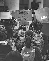 Crowd viewed from behind wearing backpacks and someone is holding a sign that reads Everybody vs Racism