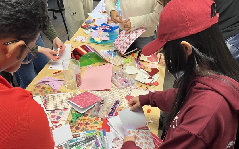 Students, mostly dressed in red, gathered at a table making Valentine's Day cards.