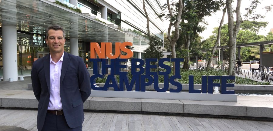 Gustavo A. Flores-Macías standing in front of a sign that reads NUS, The Best Campus Life. 
