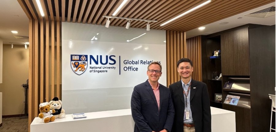 Two men standing in front of an information desk with the NUS emblem on the wall.