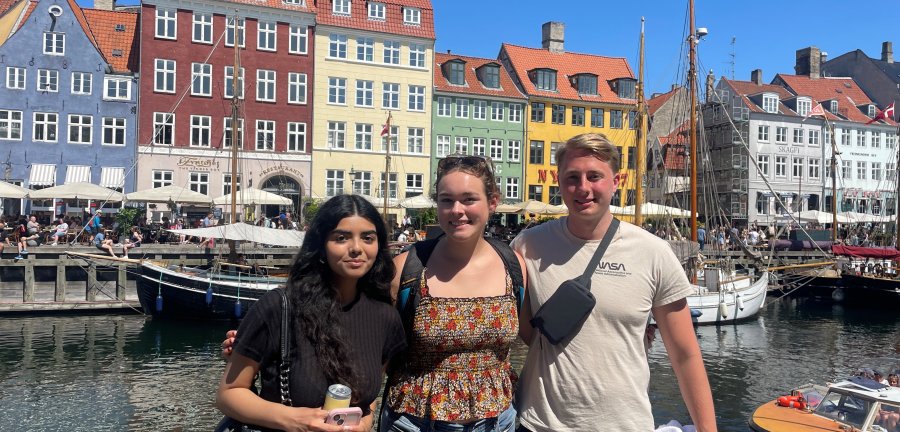 Three Cornell students smile and pose for a photo in front of the colorful buildings and canal of Nyhaven, Copenhagen.