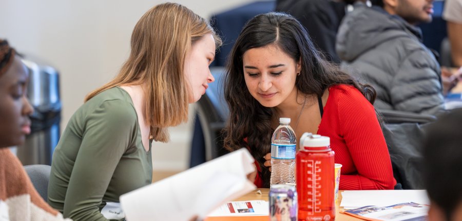 Two students conversing at a table in a room with other students around. 