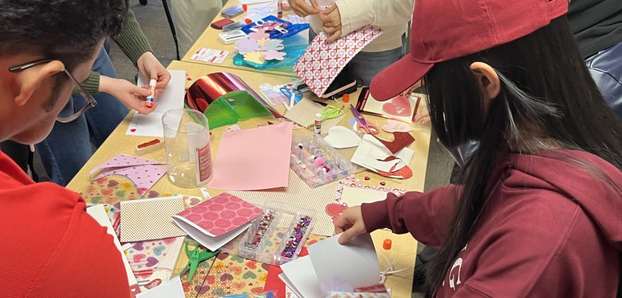 Students, mostly dressed in red, gathered at a table making Valentine's Day cards.