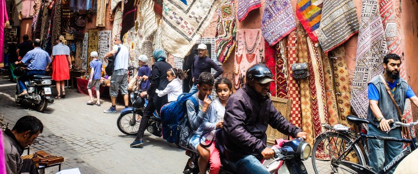 Street market with rugs hanging on display and people walking and riding bicycles.