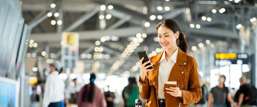 Woman standing in busy airport terminal, typing on her phone with a carry-on at her feet.