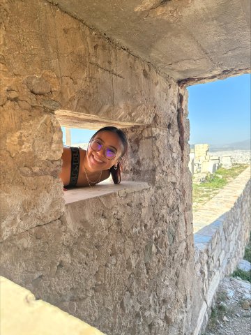 Annie Le pokes her head through the window of a stone wall in Greece.