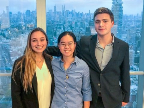 Three students with arms linked standing in front of a glass wall overlooking a city