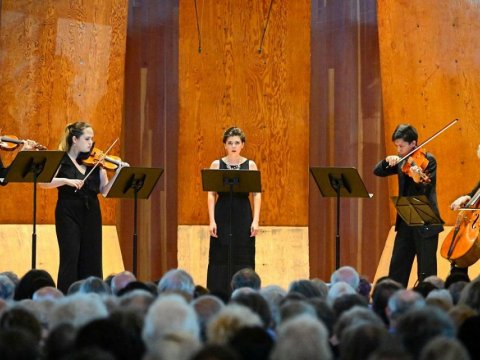 Singer standing in front of music stand with musicians playing string instruments on wither side in a concert hall
