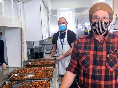 Three men wearing masks standing in a kitchen with trays of food