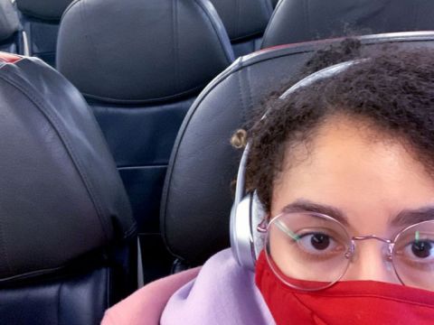 Woman sitting in a blue airline seat wearing a red face mask, surrounded by empty seats.
