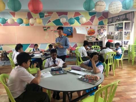 Teacher standing in classroom reading to students who are seated at round tables