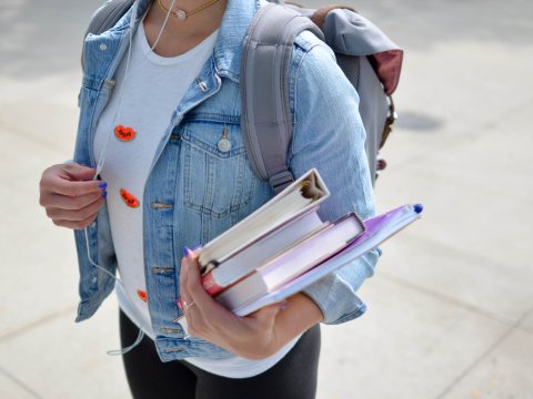 Student shown from neck down, wearing a jean jacket and backpack, carrying books