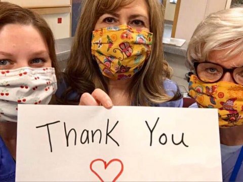 Three women healthcare professionals wearing colorful masks hold up a thank you sign.