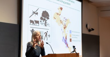 Amanda Rodewald speaks at a podium, gesturing with her hands. A large slideshow presentation behind her shows maps of bird migration.