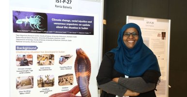 Rania Mohamed Hassan Baleela standing in front of a poster displaying her work. 