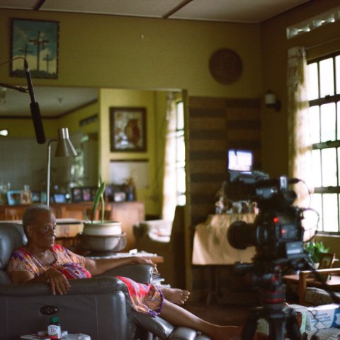 A woman sits in a leather armchair within a house. A boom mic hovers above her and a camera records her.